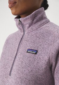 Polaire à manches longues en violet clair chiné avec un col zippé à quart, arborant un patch logo Patagonia noir sur la poitrine. Texture douce.