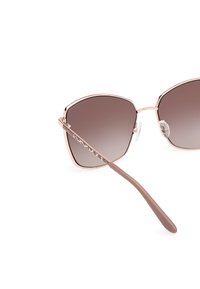 Occhiali da sole color oro rosa con una cornice pentagonale unica, lenti marroni sfumate e aste marroni testurizzate con un dettaglio del logo.