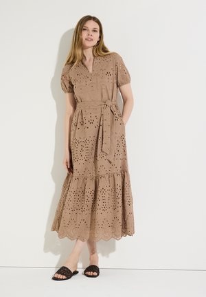 Frau trägt ein beiges Midi-Kleid mit Lochstickerei, kurzen Ärmeln, einem Taillenband und einem wellenförmigen Saum, kombiniert mit schwarzen, geflochtenen Slide-Sandalen.