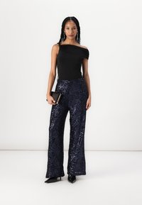 Čierny top bez ramienok v kombinácii so širokými nohavicami znavy so flitrami. Modelka drží malú čiernu kabelku a má na sebe špičaté čierne lodičky.