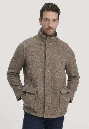 Mann in einer braunen Wolljacke mit hochgeschlossenem Kragen und Klappentaschen, der mit den Händen in den Taschen vor einem einfarbigen Hintergrund steht.