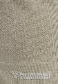 Beżowa teksturowana tkanina z ryflowanym wzorem, z białym logo na dole. Materiał wydaje się miękki i lekko elastyczny.