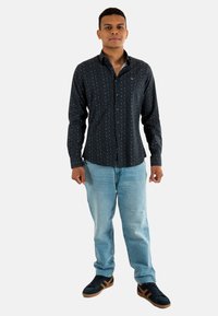 Chemise boutonnée bleu foncé avec un petit motif blanc, associée à un jean bleu clair et des baskets noires avec des accents marron.