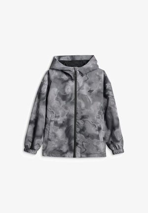 Niño de pie con las manos en los bolsillos, vistiendo una chaqueta gris con efecto tie-dye, sudadera gris, pantalones deportivos azul marino y zapatillas negras y blancas.
