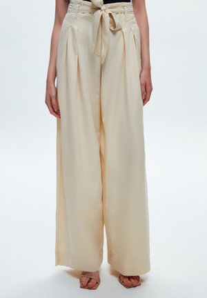 Pantaloni larghi color crema con vita legata, caratterizzati da pieghe frontali e un tessuto morbido e fluido. L'orlo è rifinito con una cucitura ordinata.