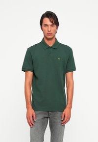 Man in een donkergroen polo T-shirt met korte mouwen en een klein geel "cK" logo, gecombineerd met vervaagde grijze jeans, staand tegen een witte achtergrond.