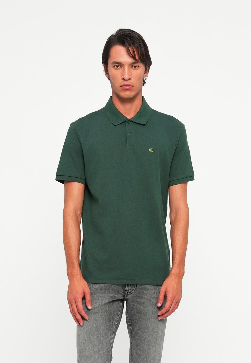 Man in een donkergroen polo T-shirt met korte mouwen en een klein geel "cK" logo, gecombineerd met vervaagde grijze jeans, staand tegen een witte achtergrond.