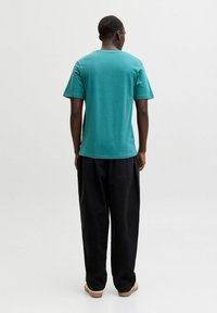 Homme debout, vu de dos, portant un t-shirt à manches courtes teal, un pantalon noir ample et des chaussures marron clair sur un fond uni.