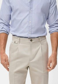 Beige broek met een rechte voorkant, knoopsluiting, decoratieve zijzak en plooi-detail; gecombineerd met een blauw-wit gestreepte shirt.