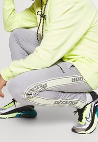 Ljust grå idrottsbyxor med kontrasterande svarta och vita ränder som har "PEGA"-tryck. Bärs med en neongul hoodie och svarta sneakers.
