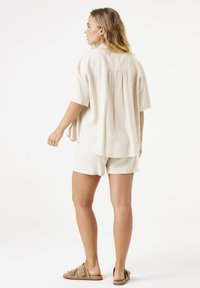 Leinenhemd mit kurzen Ärmeln und passender Shorts in Hellbeige, mit lockerem Schnitt und Rückenschlitzdetail, kombiniert mit braunen Sandalen.