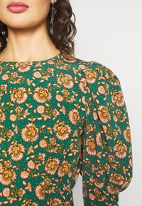 Femme portant une robe verte avec un motif floral orange et rose et des manches bouffantes aux épaules, aux cheveux foncés et bouclés, portant des boucles d'oreilles pendantes.