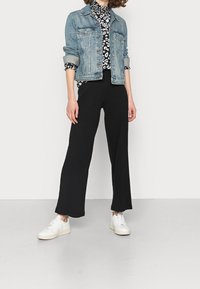Donna che indossa un giubbotto di jeans arrotolato, una blusa a righe bianco e nero, pantaloni larghi neri e sneaker bianche, in piedi su un pavimento grigio.