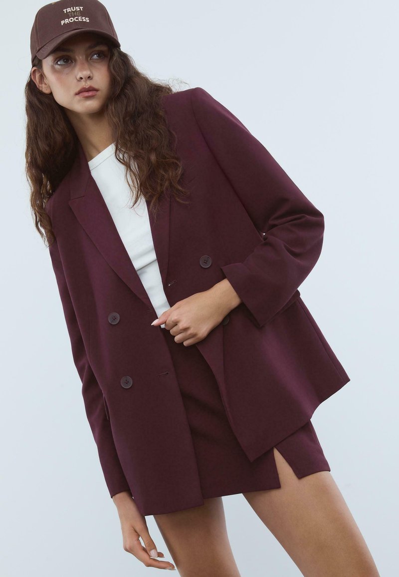 Donna con lunghi capelli ricci che indossa un blazer bordeaux doppiopetto, una gonna coordinata con uno spacco laterale, una maglietta bianca e un berretto marrone con la scritta "Fidati del Processo."