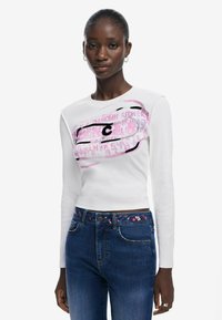 T-shirt blanc à manches longues avec un design graphique présentant un texte rose et noir. Assorti à un jean bleu foncé avec une taille brodée de fleurs.