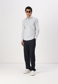Witte shirt met kleine blauwe stippen, gecombineerd met donkere broek en witte sneakers. Model met zwarte zonnebril, staande tegen een effen achtergrond.