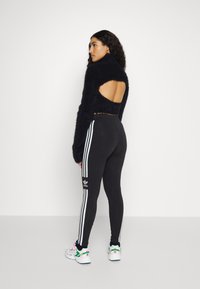 Melna pūkaina īsa džemperis ar atvērtu aizmuguri, kopā ar melniem legingiem, kuriem ir baltas trīs svītras un logs, un krāsainiem kedām.