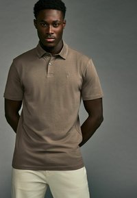 Braunes Poloshirt aus Baumwolle, mit einer Knopfleiste mit drei Knöpfen, kurzen Ärmeln und einem kleinen gestickten Logo auf der Brust.