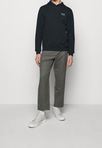 Svart huvtröja med ficka fram och logotyp, ihop med olivgrön vida byxor. Vita sneakers med grå detaljer.