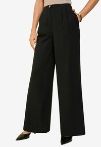 Zwarte wijde broek van soepele stof met een hoge taille, voorzien van voorplooien en een knoopsluiting. Gecombineerd met spitse zwarte schoenen.