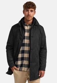 Chaqueta exterior negra con un cuello alto, que presenta botones y una cremallera, superpuesta sobre una camisa de cuadros beige y azul marino. Textura suave.