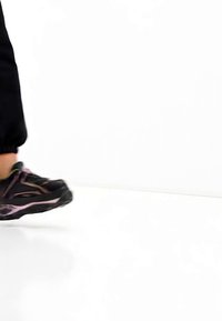 Personne avançant, portant des chaussures de sport noires avec des accents roses et un pantalon noir, sur un fond blanc.