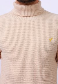 Beige coltrui met een getextureerd, wafelbreipatroon. Heeft een klein, geborduurd geel vogellogo op de linkerkant van de borst.