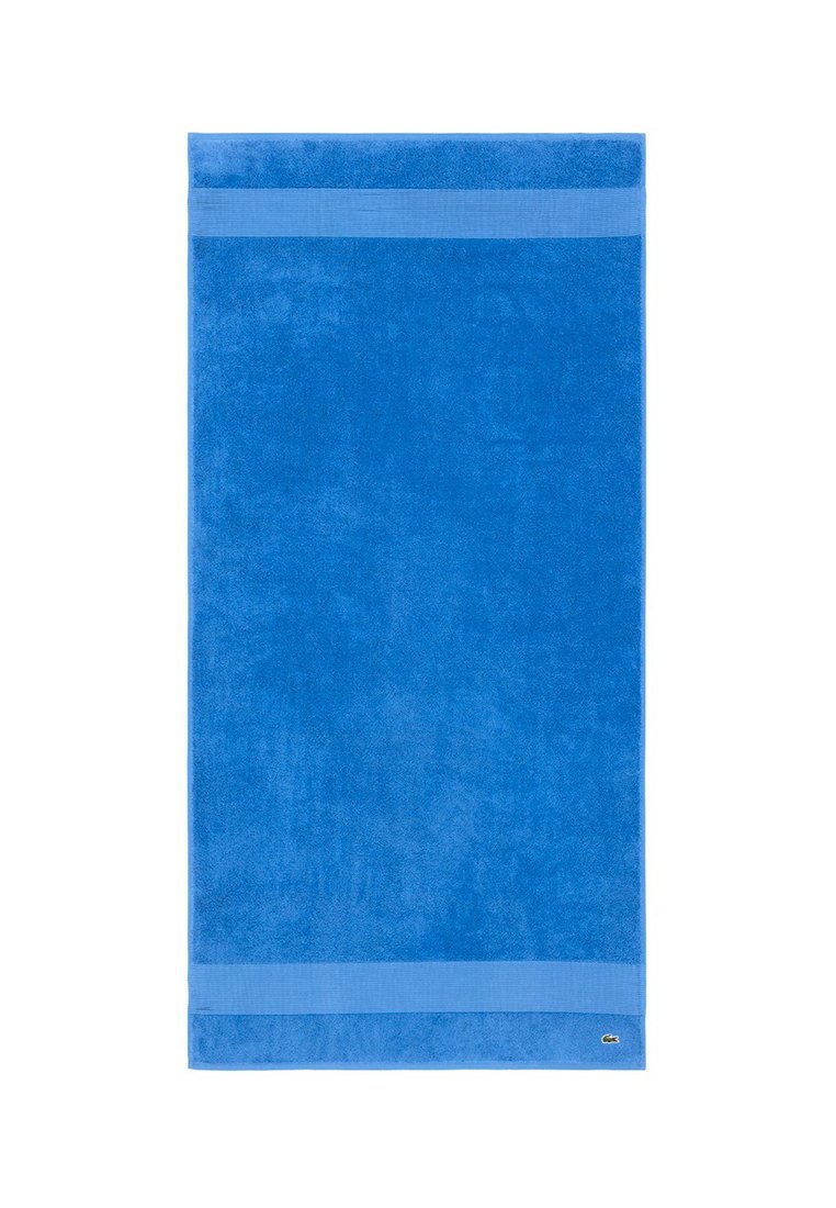 Niebieski bawełniany ręcznik kąpielowy o miękkiej fakturze z dwoma paskami jaśniejszego niebieskiego na każdym końcu. Logo Lacoste w prawym dolnym rogu.
