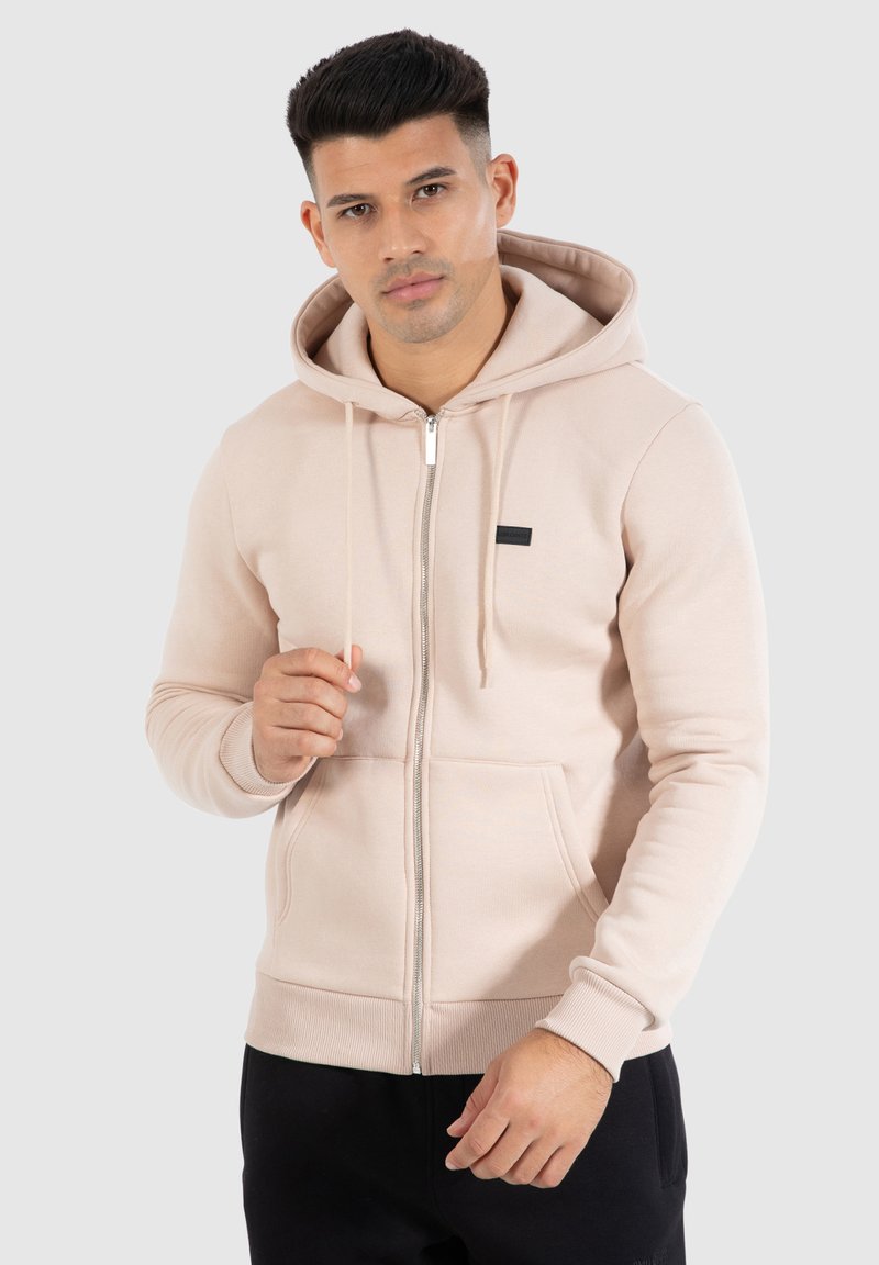 Haut zippé beige clair en tissu doux, doté d'une fermeture éclair à l'avant, de cordons de serrage et d'un petit écusson noir sur la poitrine.