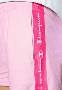 Pantalones cortos rosa claro con bolsillo lateral y una franja vertical rosa brillante que lleva el texto del logo de la marca "Champion" en blanco.