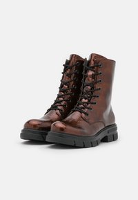 Bottes chevilles en cuir marron brillant avec lacets noirs, semelles robustes et bout renforcé. Présentent des coutures le long du bord et un détail de logo.