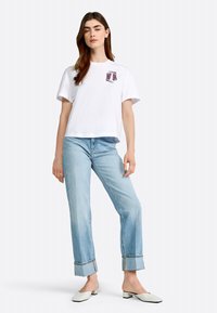 T-shirt en coton blanc, manches courtes, avec un graphique violet sur la poitrine. Assorti à un jean bleu clair et des chaussures blanches à enfiler.