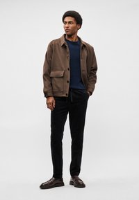 Casaco castanho com um grande bolso frontal, suéter azul-marinho, calças de veludo cotelê pretas e sapatos castanhos escuros, apresentando uma textura suave e um corte ajustado.