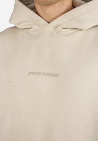 Beige hettegenser laget av myk stoff med en frontlomme og snor i hetten. "PEGADOR"-logoen subtilt trykt på brystet.