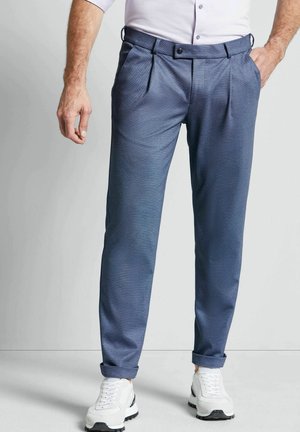Man draagt blauwe op maat gemaakte broek met omslagen, witte sneakers en een wit shirt met lange mouwen, staand tegen een effen achtergrond.