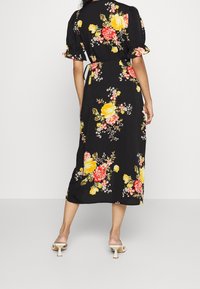 Kvinna som bär en svart midiklänning med gul och röd blommönster, puffärmar och vita klacksandaler, stående mot en enfärgad bakgrund.