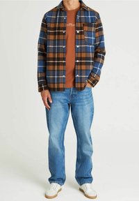 Koszula z flaneli w kratę w kolorach niebieskim, brązowym i białym, zapinana na guziki z kieszenią; pod spodem brązowy t-shirt. Jasnoniebieskie dżinsy i białe buty.