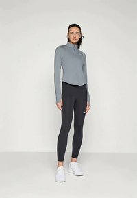 Lys grå langermet treningstopp med glidelås i kragen, kombinert med svarte tettsittende leggings. Hvite sneakers med minimale designelementer.