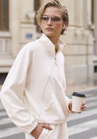 Bjela dukserica s visokim ovratnikom i zipom, s prednjim džepom. Model drži šalicu za van i nosi sunčane naočale.