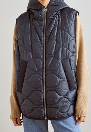 Ærmeløs sort quiltet jakke med frontlynlås, stående krave og sidelommer over beige langærmet sweater og blå jeans.