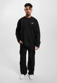 Sort langærmet skjorte med hvid "DEF" logo, kombineret med løse sorte cargo-bukser og hvide sneakers. Enkel, afslappet design.