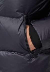 Mornarska puffer jakna s patentnim zatvaračem, s glatkom, sjajnom teksturom i rebrastim detaljem na rukavu. Tkanina ima suptilan uzorak.