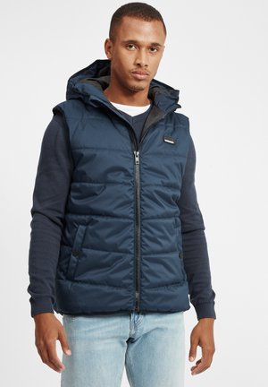 Navy blue padded vest with a hood, featuring a front zipper, side pockets, and a textured finish. Worn over a dark long-sleeve shirt.