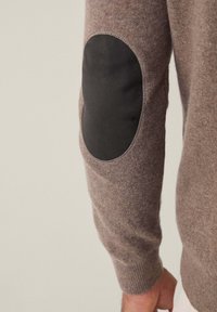 Maglione marrone lavorato a maglia con finitura texture, dotato di una toppa nera in tessuto sul gomito e polsini a costine. Vista ravvicinata di una manica.
