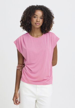 Frau mit lockigem Haar, die Creolen, ein pinkfarbenes kurzärmeliges Oberteil und weiße Hose trägt, vor einem schlichten hellen Hintergrund stehend.