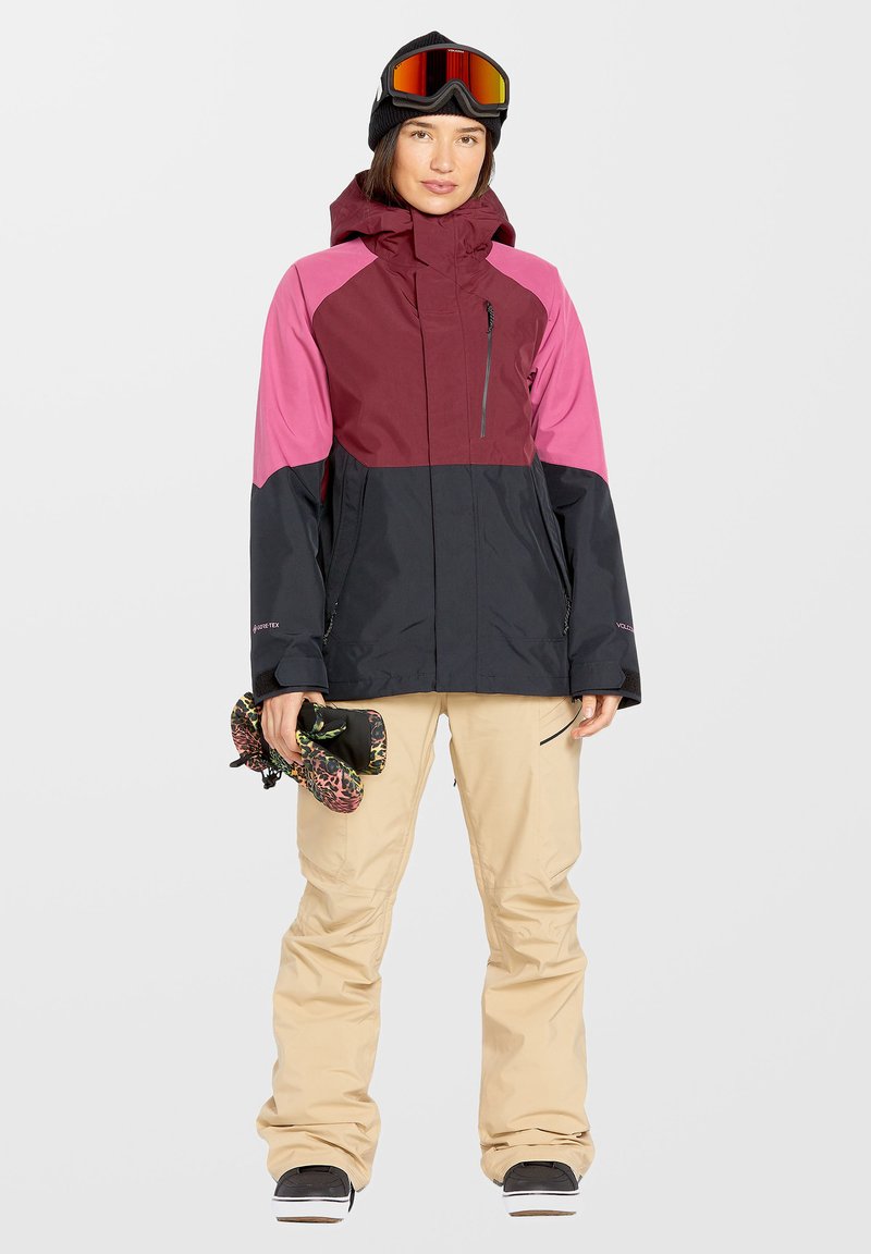 Kleurgeblokte skijas met maroon, roze en zwarte secties; ritsvak; gecombineerd met beige broeken en skihandschoenen, een donkere beanie en goggles.