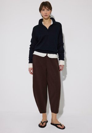 Ung person med kort hår iført en sort langærmet top, hvid skjorte under, brune bukser med vide ben og sorte sandaler, stående.