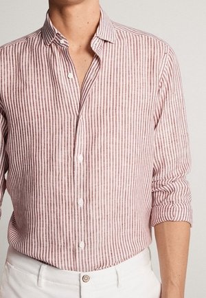 Homme portant une chemise rose clair et blanche à rayures verticales, manches longues retroussées, rentrée dans un pantalon blanc avec un bouton.