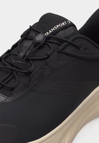 Detaljan prikaz crne sportske cipele s podesivim elastičnim vezicama, teksturiranim materijalom i bež jastučićastim potplatom, s natpisom "TRANSPORT 2" na jeziku cipele.