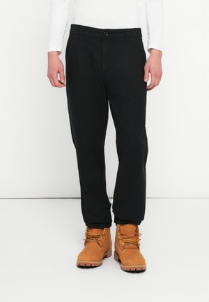 Čierne nohavice s rovno strihom, s gombíkovým zapínaním a záhybmi. Kombinované s hnedými šnurovacími topánkami a bielou košeľou s dlhým rukávom.